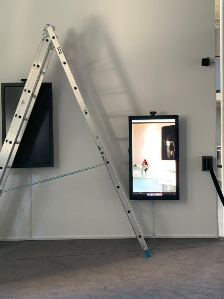 Gallery installation with a stepladder leaning against a wall between two wall-mounted screens, one dark and one showing a video of a seated figure
