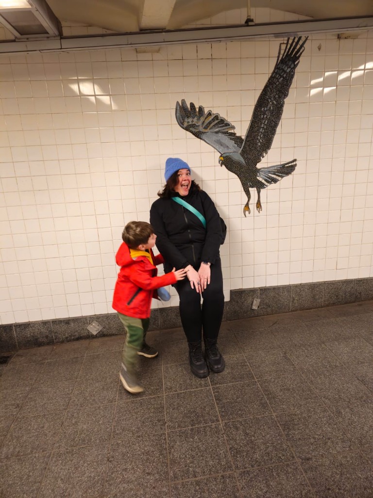 Lesley laughing with her son in a subway station, standing in front of a tiled mosaic of a hawk with outstretched wings